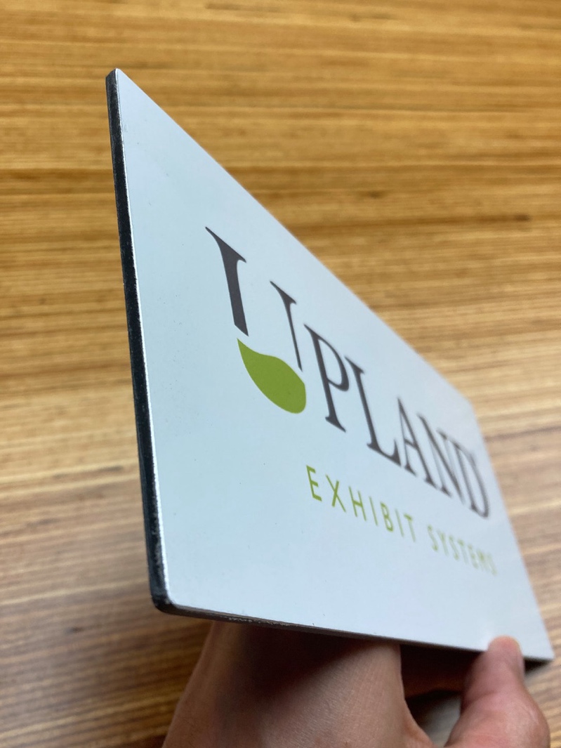A rigid ACM graphic panel held in hand, showing the laminated printed face and the cut edge