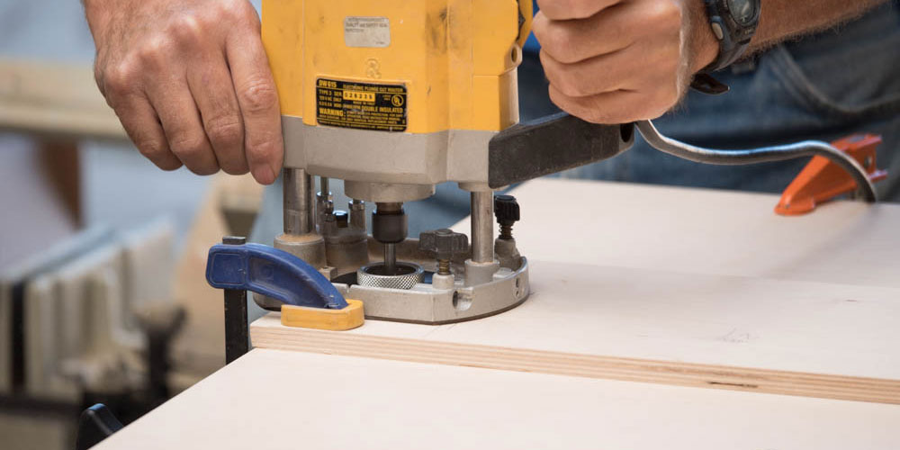 A pair of hands using an electric router to machine wood parts.